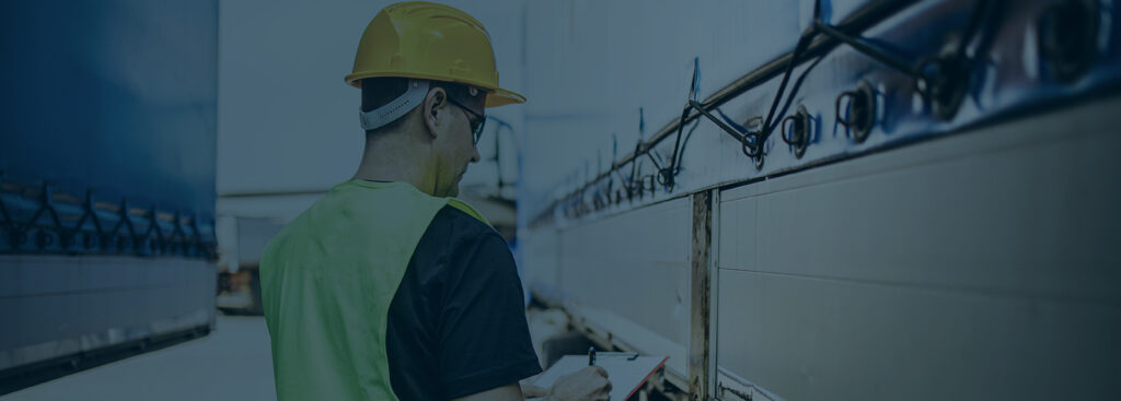 Professional male industrial truck driver with yellow protective helmet performs technical inspection of the vehicle as safety measures before next drive.